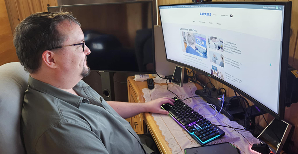 Pierre-Yves Sorel, gestionnaire de contenu à son compte chez SynergiaWeb, est assis à son bureau et travaille sur un ordinateur à écran ultra-large. Il consulte le site web Capable, qui présente des articles sur l'inclusion des personnes en situation de handicap dans le milieu professionnel. L'environnement est chaleureux, avec des murs en bois et une ambiance de bureau à domicile.