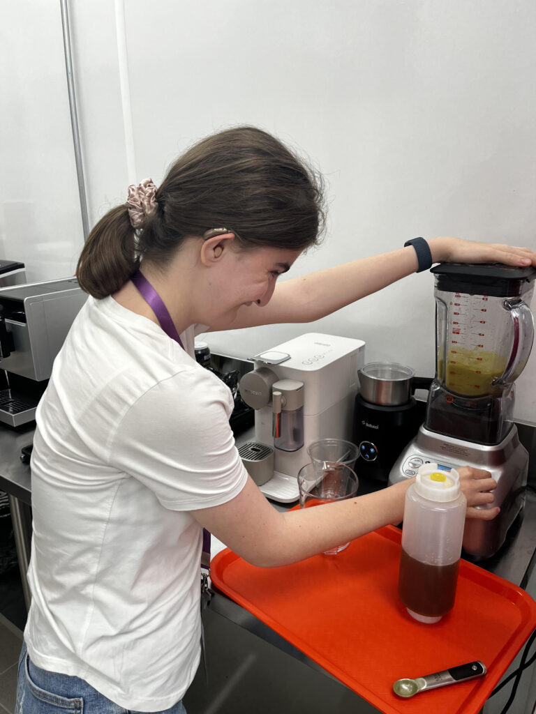 Une jeune femme avec une déficience intellectuelle est concentrée et souriante en utilisant un mélangeur dans une cuisine professionnelle. Elle porte un appareil auditif, une montre intelligente et une queue de cheval avec un chouchou rose. Elle prépare une boisson à côté d’une machine à café, sur un plateau orange. Cette jeune femme est intégrée dans l’entreprise CitiPouss, dirigée par sa mère Marianne Faraud.